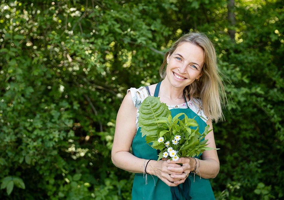 Lächelnde Frau mit Schürze hält draußen verschiedene Kräuter und Pflanzen in ihrer Hand.